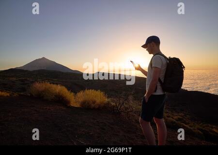 Hiker using mobile phone on hill above clouds at sunset. Young man with backpack against landscape. Colcano Pico de Teide in Canary Islands, Spain. Stock Photo