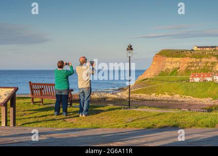 A couple take photographs on smart phones on a cliff overlooking a harbour. Cottages are visible in the distance and an evening light throws long shad Stock Photo