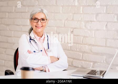 Portrait shot of senior female doctor sitting at desk behind her laptop while working. Stock Photo