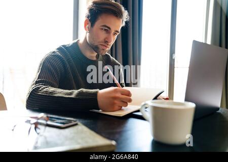 Serious mid aged man working on laptop computer from home, taking notes Stock Photo