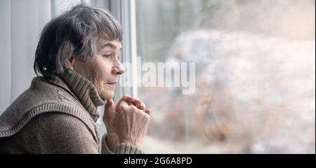 An elderly well-groomed woman, a pensioner, looks thoughtfully from the window  Stock Photo