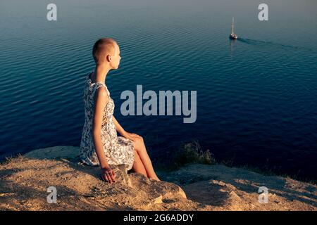 teenage girl in short hair dress sits on a cliff over a sea on a sunny day, looking at the ship Stock Photo