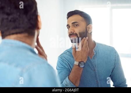 Curious thoughtful indian man touching beard on face looking at mirror Stock Photo
