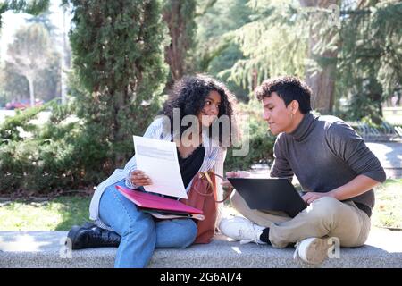 Two latin students studying together sitting on a bench outdoors. University life at campus. Stock Photo