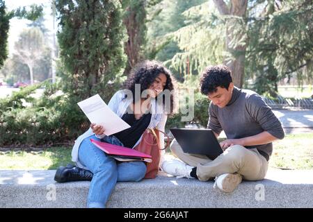 Two latin students smiling studying together sitting on a bench outdoors. University life at campus. Stock Photo