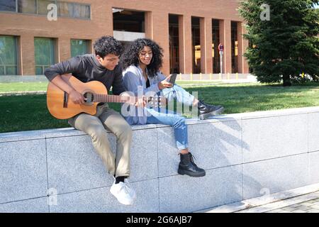 Young latin couple playing the guitar and using the smartphone at the university campus. University life, millennial generation. Stock Photo