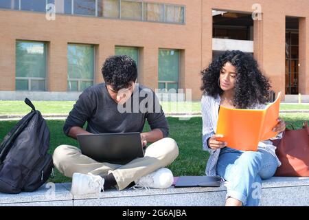 Two latin students studying from their lecture notes sitting on a wall outdoors. University life at campus. Stock Photo