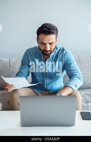 Serious indian man with paper looking at laptop screen checking on investments Stock Photo