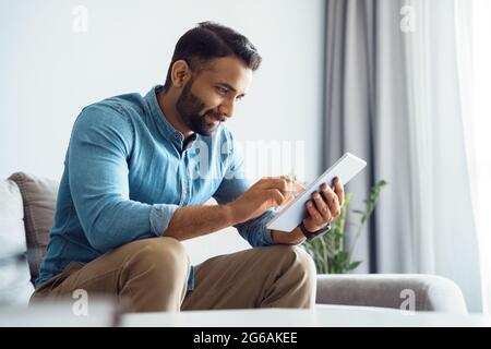 Millennial indian man using tablet relaxing on couch sofa in living room Stock Photo