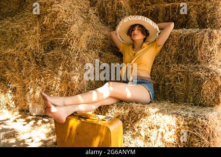 Portrait of a woman with a straw hat, yellow shirt, jean shorts and a yellow suitcase sitting in a hayloft. Stock Photo