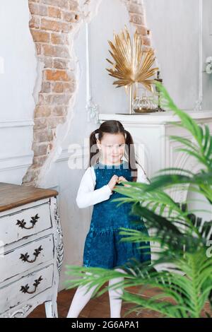 a full-length portrait of a beautiful five-year-old girl in a denim dress making a heart symbol with her fingers up in a home environment with potted Stock Photo
