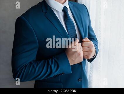A businessman in a white shirt and tie adjusts the blue suit standing at the window. Stock Photo