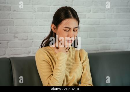 Young Asian woman at home sitting on couch resting and touching neck due to sore throat with eyes closed and feeling discomfort Stock Photo