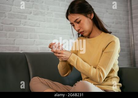 Young Asian upset woman holding hand and pressing points in palm while suffering from wrist pain sitting on sofa at home Stock Photo