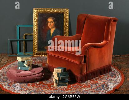 Red vintage armchair placed on ornamental carpet near pouf and stacks of aged books against frames with classic portrait Stock Photo