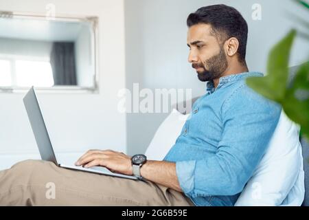 Serious indian business man executive working on laptop sitting on home bed Stock Photo