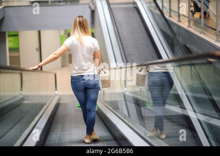 Young woman in white T-shirt and jeans on escalator in shopping center. Shopping and pleasure. Lifestyle. Everyday life in city. Goods and consumption Stock Photo