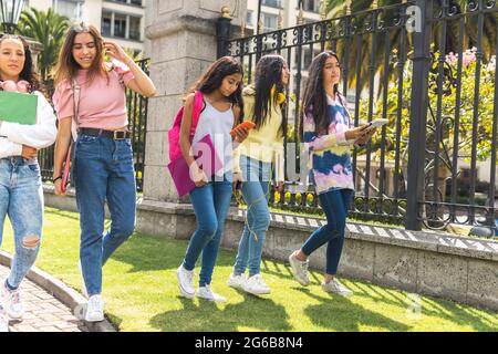 Group of Latina teen friends students walking with their backpacks, notebooks, laptop and cell phone on their way to high school Stock Photo