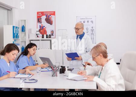 Physiotherapist senior man discussing sickness examination with medical team analyzing disease symptoms presenting healthcare treatment. Practitioners doctors working in conference meeting room Stock Photo