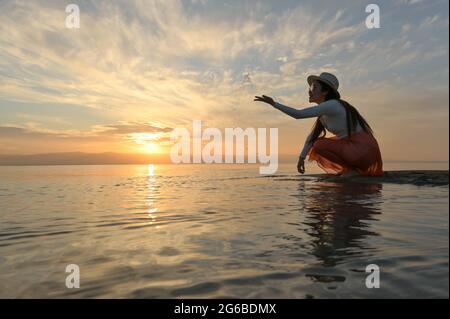 Beautiful woman crouching by sea throwing water in the air at sunset, Thailand Stock Photo