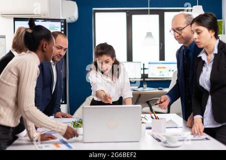 Diverse team of workers talking, looking at documents and analyzing graphs data standing at conference desk. Multiethnic colleagues working planning success financial strategy discussing in office Stock Photo