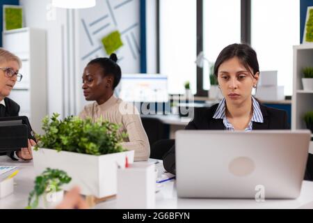 Concentrated manager typing on laptop sitting at desk in start up business office. Diverse colleagues working in background. Multiethnic coworkers planning new financial project. Stock Photo