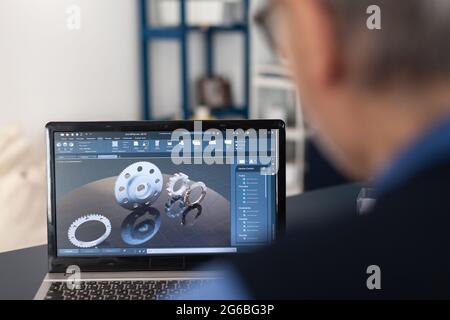 Engineer designed gears parts using professional software. Experienced engineer working from home office on mechanical project using laptop computer technology and wife is reading a book on sofa. Stock Photo