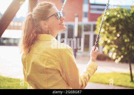 Happy young positive woman on swing in sunset, profile view, outdoors in park Stock Photo