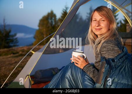 Portrait of young traveler woman with tender smile on face, covered by sleeping bag with cup of tea in her hands relaxing in tent on fresh mountain air. Concept of hiking, traveling and camping. Stock Photo