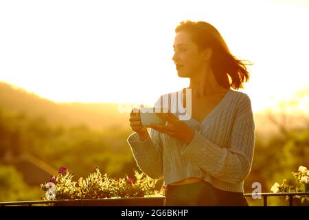 Confident woman holding coffee cup contemplating at sunset in a balcony Stock Photo
