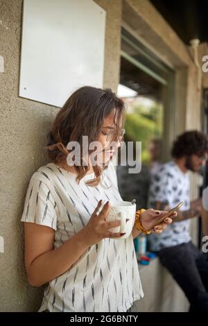 A young woman is enjoying coffee and using smartphone during a break in a relaxed atmosphere outside the office Stock Photo