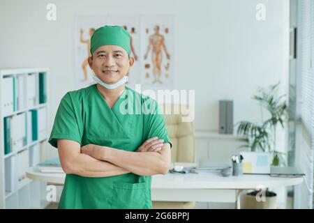 Smiling experienced surgeon in green scrubs and medical mask standing in office of clinic and looking at camera Stock Photo