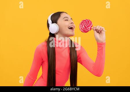 happy kid singer listen music in headphones singing with lollypop on yellow background, singer Stock Photo