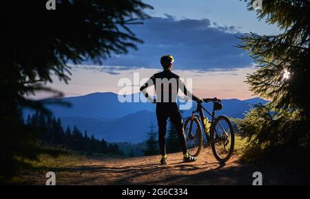 Back view on male cyclist standing holding his bike, watching beautiful landscape, mountains peaks and sunset, surrounded with spruces in the evening. Active lifestyle concept Stock Photo