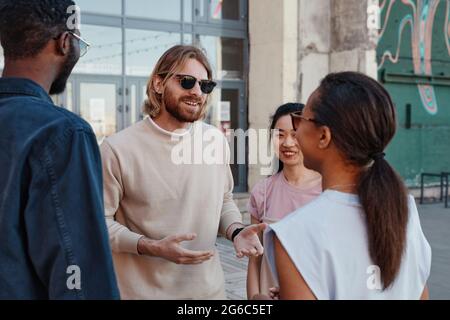 Diverse group of contemporary young people chatting outdoors in urban city setting, focus on smiling man wearing sunglasses Stock Photo