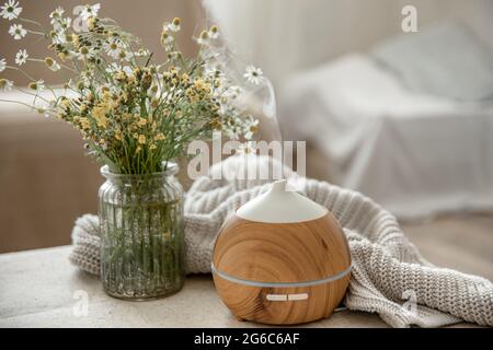 Modern oil aroma diffuser in the living room on the table with knitted element and flowers. Stock Photo