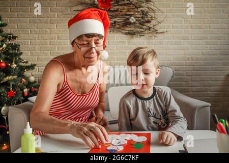 A Little boy with grandmother make Christmas craft, greeting card. Christmas tree on background. Stock Photo