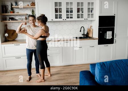 Young happy caucasian spouses, couple staying in kitchen at home. Attractive family of two doing morning exercises, having fun together, showing Stock Photo