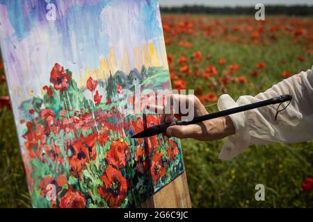 Hand with brush close-up. Mature woman artist paints a picture on a field of blooming poppies Stock Photo