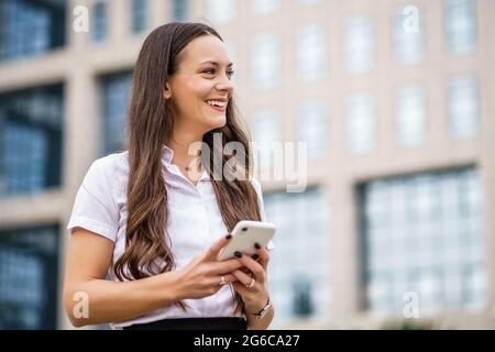 Portrait of happy businesswoman typing on smartphone in front of company building. Stock Photo
