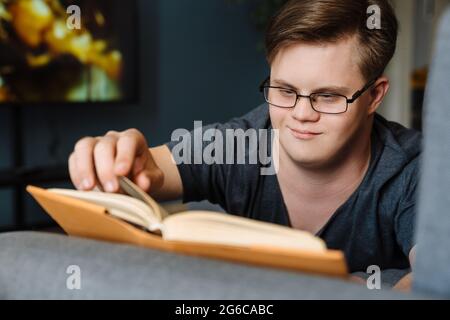 Young man with down syndrome reading book while lying on couch at home Stock Photo