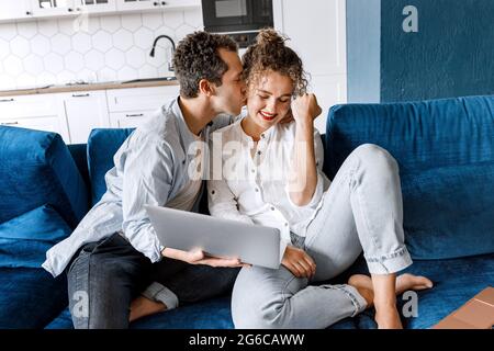 Millennial joyful couple sitting on sofa and kissing,using laptop together doing online shopping for home, surfing web, checking social media Stock Photo