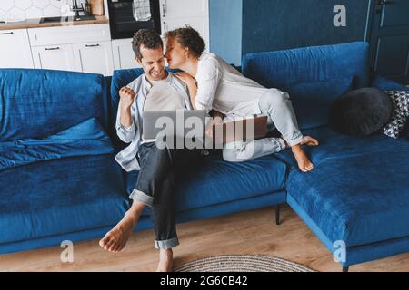 Young freelance couple working online with laptops together at home, sitting on sofa. Handsome overjoyed male celebrating victory, success and his Stock Photo