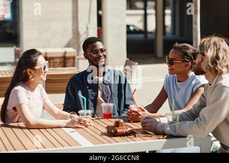 Diverse group of young people enjoying cocktails while sitting at cafe table outdoors lit by sunlight Stock Photo