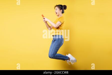 Full body profile photo of young asian girl jumping high holding a phone writing a new social media post, isolated on blue background Stock Photo