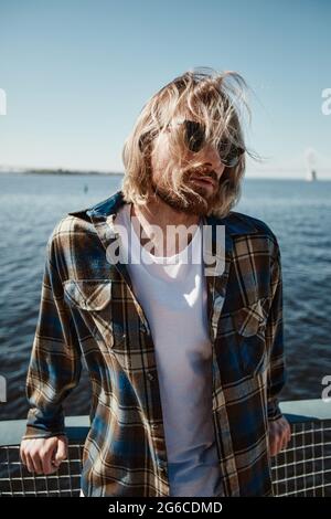 Vertical film portrait of long haired young man wearing sunglasses while posing by river in wind Stock Photo