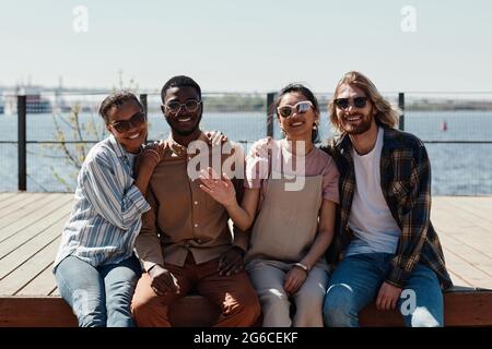 Diverse group of young people smiling at camera while posing outdoors by river, all wearing sunglasses Stock Photo