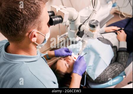 Close up of male dentist in medical mask using dental diagnostic microscope while checking patient teeth. Young woman lying in dental chair at dental office. Concept of dentistry and dental care. Stock Photo
