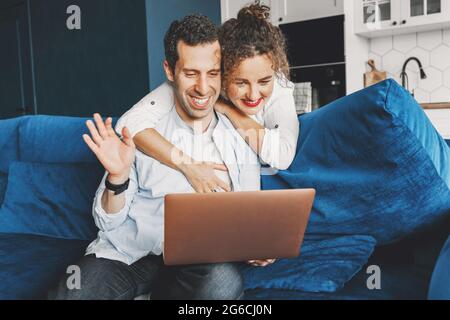 Happy millennial man use laptop at home on sofa in living room waving hello to friends and family remotely during quarantine. Joyful smiling woman Stock Photo