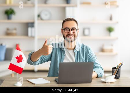Happy young guy sitting at table with flag of Canada, using laptop computer, showing thumb up gesture at home Stock Photo